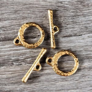 May include: Three gold-toned metal toggle clasps with a hammered texture. The clasps are in the shape of a circle and a bar.
