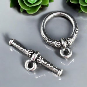 May include: Silver toggle clasp with a decorative design. The clasp has a round ring and a bar with a small loop at the end.