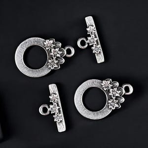 May include: Four silver-toned toggle clasps with floral accents. Two clasps are circular with a floral cluster, and two are bar-shaped with floral details. The clasps are on a black background.