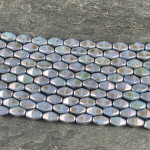 Pode incluir: Close-up de contas iridescentes em forma oval, em tons de azul, prata e dourado. As contas t&ecirc;m um brilho met&aacute;lico e est&atilde;o dispostas em um padr&atilde;o de favo de mel. Estas contas s&atilde;o adequadas para fazer joias.