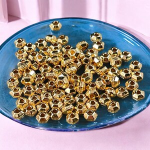 May include: A collection of small, gold-colored, geometric beads. The beads have a multifaceted design and are scattered on a blue glass plate. The beads are shiny and reflect light, creating a visually appealing display. These beads are suitable for jewelry making and crafts.
