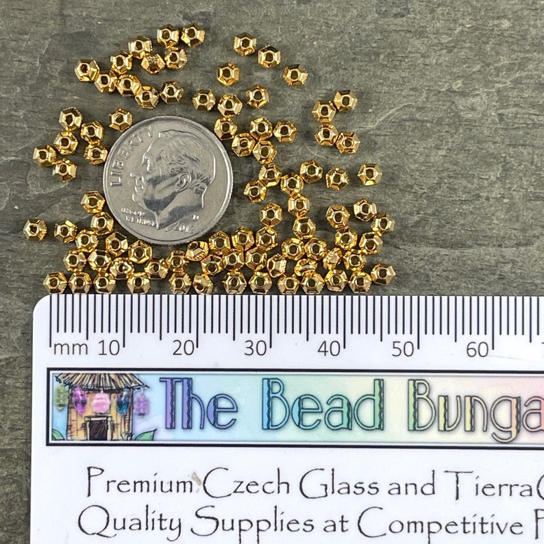 May include: A collection of small, gold-colored, faceted beads scattered on a gray surface. A U.S. dime is included for size reference. A ruler with millimeter markings is visible. The image also includes text that reads "The Bead Bungalow".