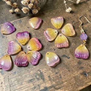May include: A collection of translucent, petal-shaped beads in shades of yellow, pink, and purple, scattered on a wooden surface. One earring with a gold hook and purple bead is also visible. The beads have a hole for stringing.