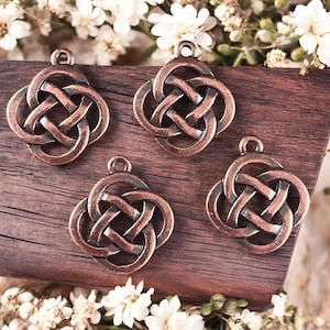 May include: Four bronze-colored Celtic knot charms with a loop at the top for hanging. The charms are displayed on a dark wooden surface, with white flowers in the background. The charms have an intricate interwoven design.