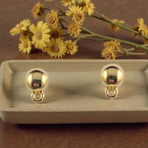 May include: A pair of gold-colored clip-on earrings with a spherical design. The earrings are displayed on a light brown wooden tray, with yellow flowers in the background. The earrings have a shiny, reflective surface.