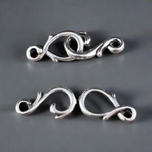 May include: Two silver-toned metal clasps with a decorative, ornate design. The clasps are shaped like a stylised vine or branch.