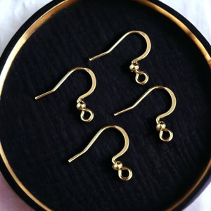 May include: Four gold-toned metal earring hooks, each featuring a curved wire design, a small decorative bead, and a circular loop at the bottom for attaching jewelry components. These earring findings are arranged in a cluster on a dark surface.