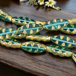 May include: Close-up of a strand of artisan beads. The beads are oval-shaped with a teal center and gold-colored edges. The beads are arranged on a dark wooden surface, with a few small white flowers in the background.