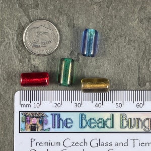 May include: Four glass beads in different colors: red, green, blue, and gold. The beads are approximately 1 cm long and are arranged on a ruler with millimeter markings.