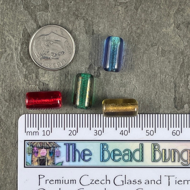 May include: Four glass beads in different colors: red, green, blue, and gold. The beads are approximately 1 cm long and are arranged on a ruler with millimeter markings.