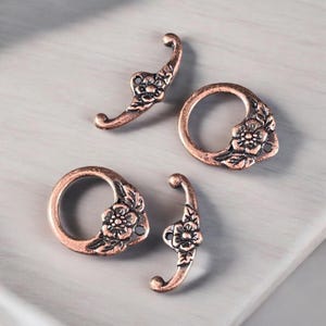 May include: Four antique copper-colored metal jewelry findings. Two are curved with floral details, and two are circular with floral accents. The findings are arranged on a light gray surface.