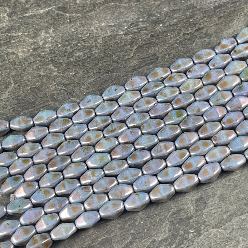 Pode incluir: Close-up de fileiras de contas iridescentes em forma oval, em tons de azul, castanho e prata. As contas t&ecirc;m um brilho met&aacute;lico e s&atilde;o enfiadas, perfeitas para a cria&ccedil;&atilde;o de joias. As contas est&atilde;o dispostas diagonalmente.
