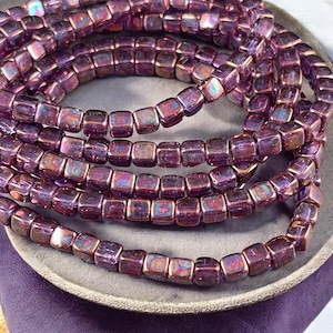May include: A strand of iridescent purple square glass beads. The beads have a metallic sheen and are arranged in a circular pattern.