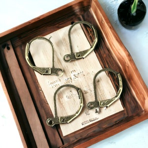 May include: Four gold-toned metal clasps or earring hooks in a wooden tray. The clasps are arranged in a pattern on a light brown surface.