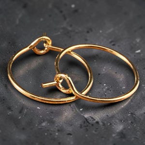 May include: A pair of gold-toned hoop earrings featuring a thin, circular wire design. Each earring has a simple latch mechanism with a small post that secures into a loop, creating a continuous, minimalist circular shape for the ear.
