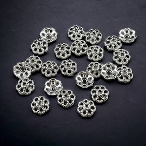 May include: A group of silver-toned metal bead caps with a decorative flower design. The caps are scattered on a dark grey surface.