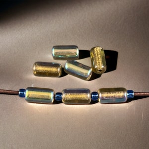 May include: A close-up of iridescent gold glass beads with a textured surface. The beads are strung on a brown cord with blue spacer beads.