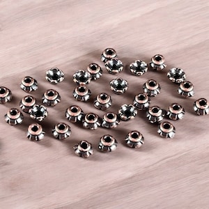 May include: A collection of small, flower-shaped metal beads with a dark, antiqued copper finish. Each bead features a central hole and a scalloped edge, creating a petal-like appearance. The beads are scattered across the surface.