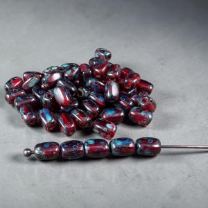 May include: A collection of barrel-shaped beads featuring a deep red base color with mottled blue and black accents. A single metal pin holds five of these beads in a horizontal row, while a pile of identical loose beads sits behind them.