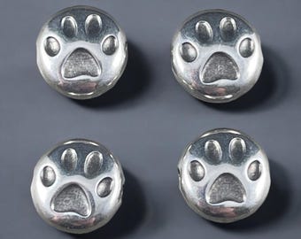 May include: Four circular silver-toned beads, each featuring a recessed paw print design on the front surface. The beads have a smooth, polished finish with a contrasting matte texture within the stamped paw print indentations.