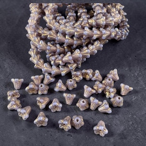 May include: A collection of small, bell-shaped beads with a pale lavender base and a metallic gold finish on the textured, fluted exterior. The beads are arranged in a loose pile, with some strung together in a long, coiled strand.