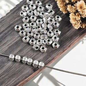 May include: A collection of silver-colored, geometric beads with multiple facets and hexagonal holes. Some beads are strung on a thin metal wire, while others are scattered on a wooden surface. Dried flowers are in the background.