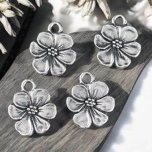 May include: Four silver-toned flower charms with a detailed petal design and a small loop at the top for stringing. The charms are arranged on a dark, textured surface, suggesting a craft or jewelry-making theme.