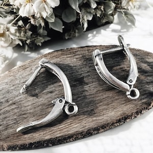 May include: A pair of silver-toned earrings with a unique, curved design. Each earring features a hinged closure and a small loop at the bottom. The earrings are displayed on a wooden surface, with white flowers in the background.