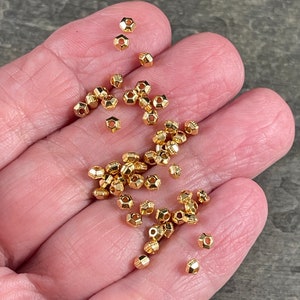 May include: A collection of small, gold-colored, faceted beads. The beads have a hexagonal shape with multiple facets, creating a reflective surface. They are scattered across a neutral background, suggesting their use in jewelry or craft projects.
