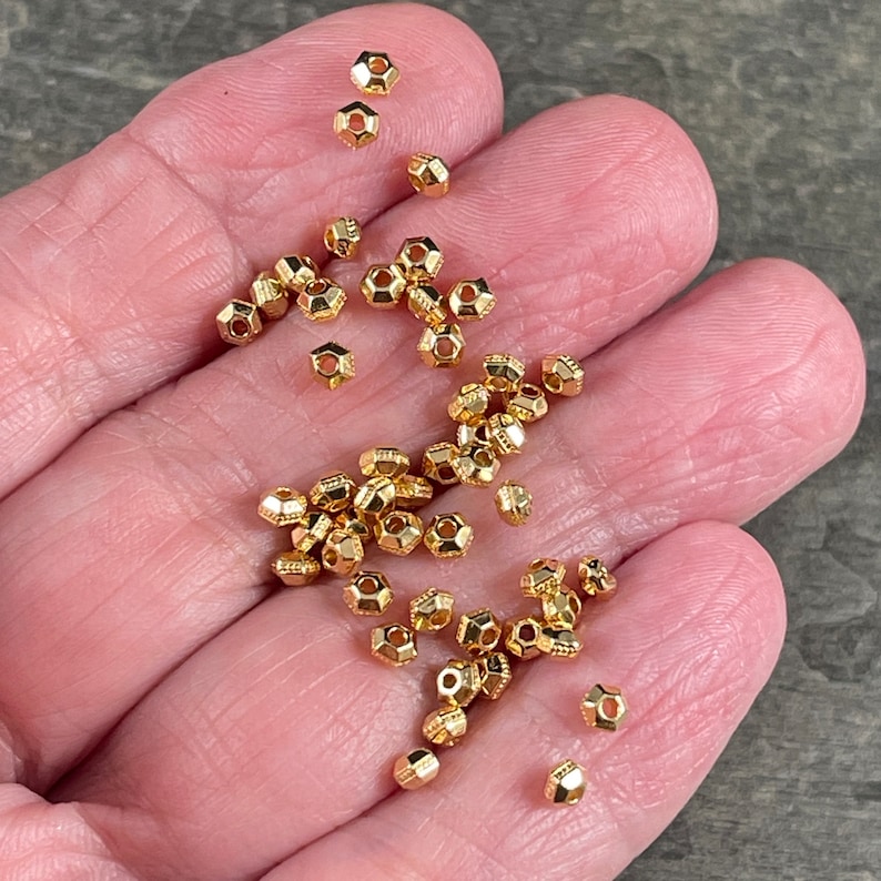 May include: A collection of small, gold-colored, faceted beads. The beads have a hexagonal shape with multiple facets, creating a reflective surface. They are scattered across a neutral background, suggesting their use in jewelry or craft projects.