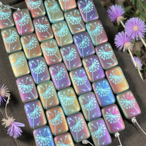 May include: Rectangular iridescent beads with a dandelion design in light blue. The beads have a metallic sheen, shifting between gold, pink, and purple hues. The beads are strung together, and purple flowers are in the background.