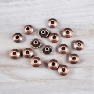 May include: A collection of copper-colored metal beads with a central hole. The beads have a slightly domed shape and a dark, recessed center. They are scattered on a light wood-grain surface, creating a textured and tactile appearance.