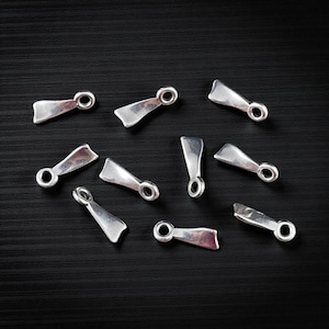May include: A collection of silver-colored metal findings, each with a paddle-shaped body and a circular loop at one end. The findings are scattered on a dark, textured surface, creating a visually appealing arrangement. These findings are likely used in jewelry making or crafting.