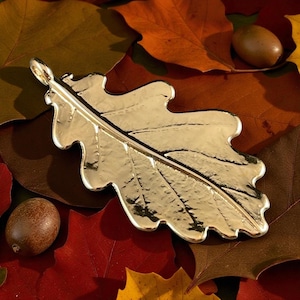 May include: A gold-coloured oak leaf pendant with detailed features, set on a bed of autumnal leaves in various colours. Two acorns are also visible. A nature-themed accessory.