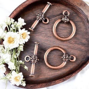 May include: Four antique copper-toned metal toggle clasps with decorative bead accents. Two are round with a loop on top, and two are rectangular with a loop on one end.