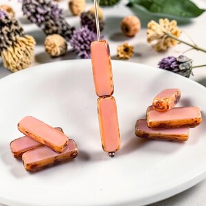 May include: Close-up of pink and brown rectangular beads. Some beads are strung on a wire, while others are scattered on a white plate. The beads have a translucent pink center with brown edges. Dried flowers are in the background.