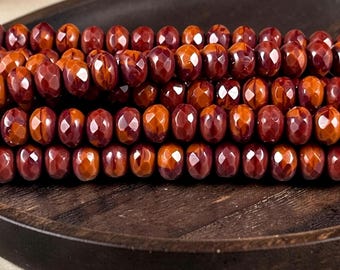 May include: A collection of faceted, round beads in shades of deep red, burnt orange, and brown. The beads are arranged in multiple horizontal rows, showcasing a polished, reflective surface with geometric facets that catch the light.