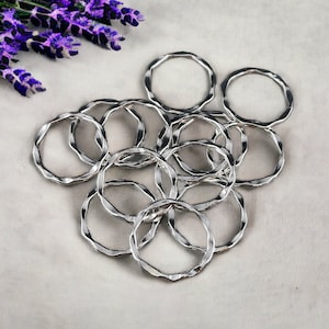 May include: A collection of shiny silver metal rings with a wavy, textured design. The rings are arranged in a cluster on a light gray surface, with a sprig of purple flowers in the upper left corner. These rings could be used for crafts or jewelry making.