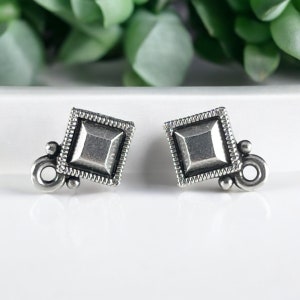 May include: A pair of silver-toned square stud earrings. Each earring features a raised square center, surrounded by a beaded border. Decorative elements include small spheres and a circular loop for attachment. The earrings are displayed on a white surface.