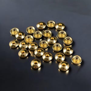 May include: A scattering of small, shiny, gold-coloured metal rivets. Each rivet has a rounded top with a central hole and a flat base. The rivets are spread across a dark, reflective surface.
