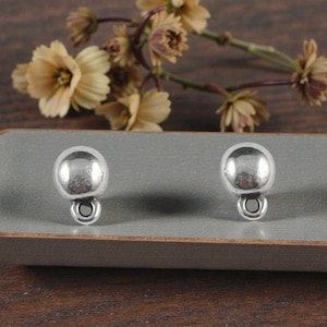 May include: Two silver-colored, round earring components with a small loop at the bottom. The earrings are displayed on a gray surface, with blurred flowers in the background. The earrings have a shiny, reflective surface.