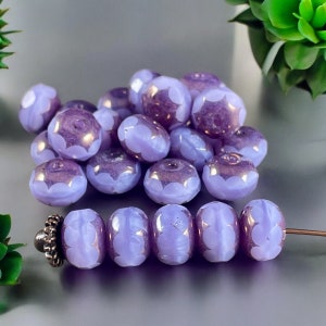 May include: A collection of round, lavender-colored glass beads featuring a scalloped, petal-like pattern in a lighter shade of purple. Some beads are loose in a pile, while five identical beads are strung on a thin metal wire with a silver end cap.