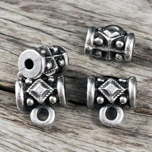 May include: Close-up of several silver-toned metal beads. Each bead features a geometric design with a central diamond shape, surrounded by smaller circular accents. The beads have a cylindrical shape with a hole for stringing and a loop at the bottom.