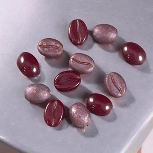 May include: A collection of coffee bean-shaped beads in burgundy and lavender hues. Some beads are glossy, others have a matte texture. The beads are arranged on a light grey surface. Ideal for jewellery making.