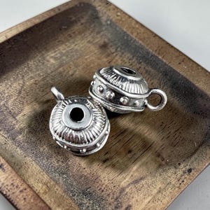 May include: Two silver-colored, ornate beads with a cup-like design. Each bead features a central hole, a small loop, and decorative detailing. The beads are displayed on a wooden surface, showcasing their intricate craftsmanship.