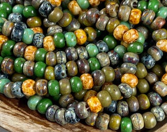 May include: A close-up of a collection of colorful glass beads. The beads are a mix of green, brown, and orange colors with a speckled pattern. The beads are arranged in a pile, showing their various shapes and sizes.