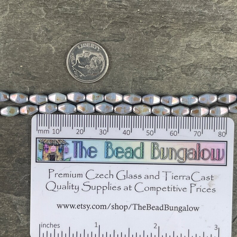 Pode incluir: Fileiras de contas de vidro checo azul claro iridescente com uma moeda de prata para escala. As contas t&ecirc;m aproximadamente 0,6 cm de comprimento. Uma r&eacute;gua e um cart&atilde;o de visita para The Bead Bungalow tamb&eacute;m est&atilde;o na imagem.