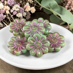 May include: Six green flower-shaped beads with pink accents are arranged on a white plate. The beads have a glossy finish and a detailed floral design, perfect for jewelry making or crafting projects.