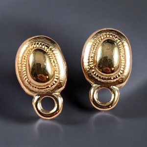 May include: Two gold-colored oval-shaped jewelry findings with a loop at the bottom. The findings have a textured border and a smooth, raised center. The findings are likely for earrings or pendants.