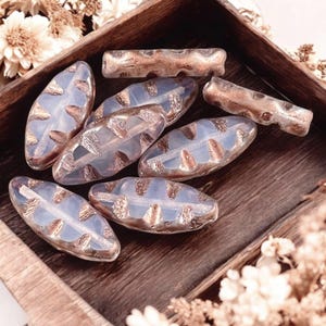 May include: A collection of elongated, oval-shaped glass beads featuring a translucent, milky white base with metallic copper-colored textured accents along the center and edges. The beads have a smooth, polished finish and a distinct, segmented design.